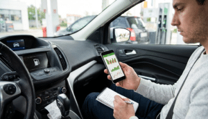 Vue de l'intérieur d'une voiture garée dans une station-service. Un homme assis côté conducteur tient un smartphone affichant des graphiques de consommation de carburant de couleur verte et rouge. De l'autre main, il écrit des notes dans un carnet posé sur ses genoux. Sur la console centrale, l'écran de bord de la voiture affiche une icône de pompe à essence, et un ticket de caisse est visible près du levier de vitesse. En arrière-plan, à travers les vitres, on distingue des pompes à carburant et d'autres véhicules.